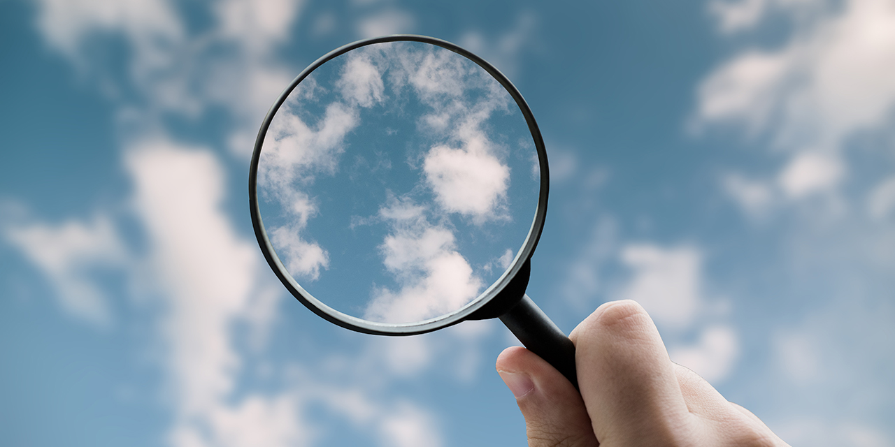 Hand holding a magnifying glass against a blue sky with scattered white clouds.