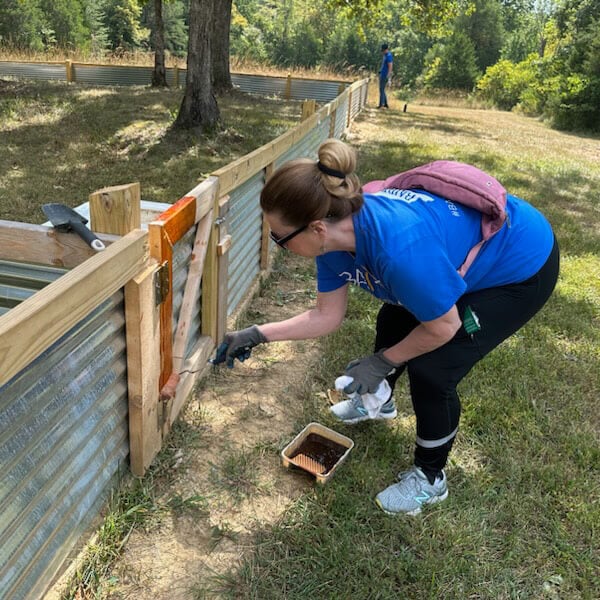 An associate wearing a Baird Gives Back t-shirt stains the wood on a fence