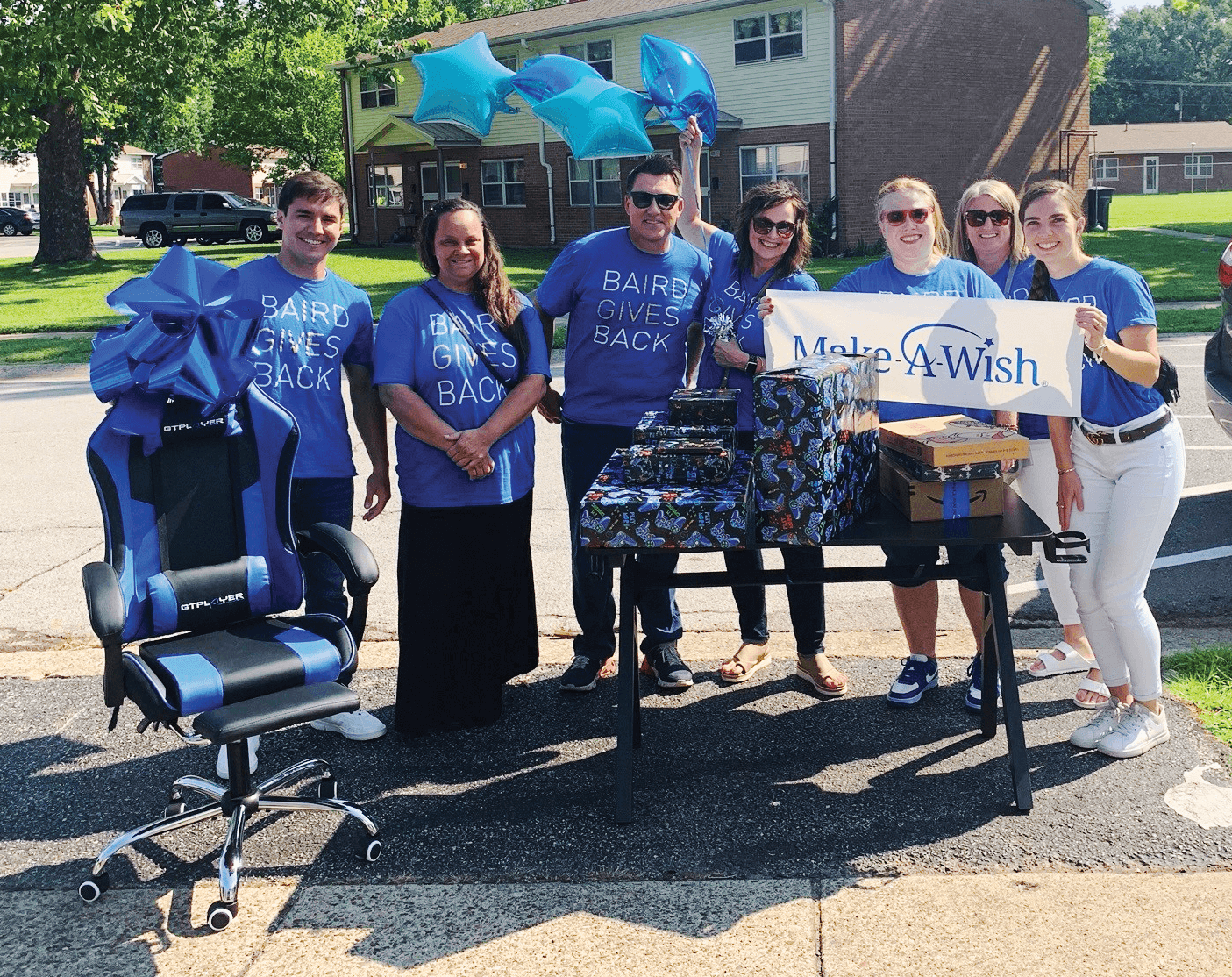 A group of Baird associates stand with a gaming chair, giftwrapped presents, and a sign for the Make a Wish foundation