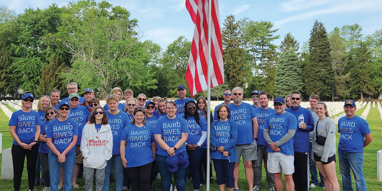 A large group of Baird associates in Baird Gives Back t-shirts stand behind an American flag at Zachary Taylor cemetery